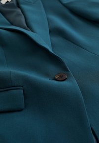 Turkos kavaj i strukturerat tyg, med en svart knapp, spetsiga slag och en vänster bröstficka. Skräddarsydd design med rena linjer.