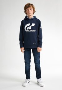 Marinblå hoodie med en vit "Gran Turismo"-logotyp och grå detaljer, ihop med blå jeans och vita sneakers. Enkelt, avslappnat design.