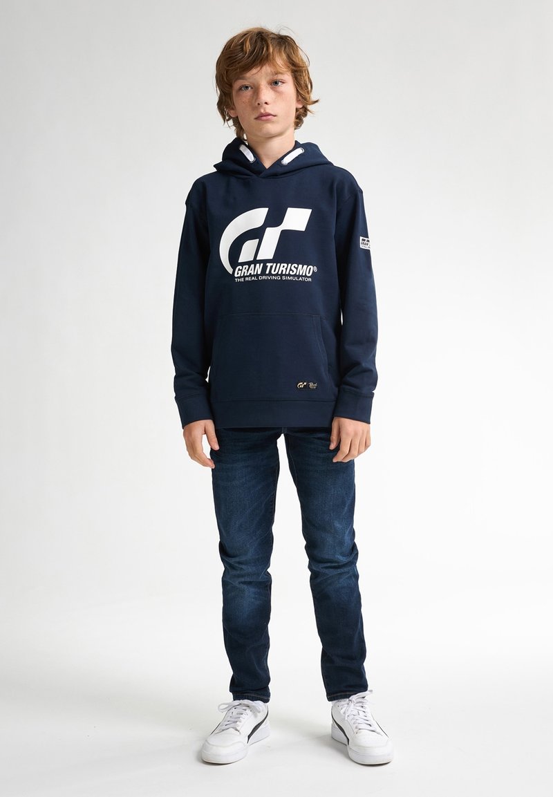 Marinblå hoodie med en vit "Gran Turismo"-logotyp och grå detaljer, ihop med blå jeans och vita sneakers. Enkelt, avslappnat design.
