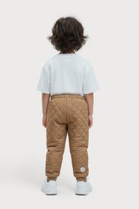 Barn med bølgende brunt hår iført en simpel hvid T-shirt, quiltede brune bukser og hvide sneakers, som står med ryggen til på en ensfarvet baggrund.