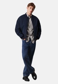 Giacca bomber navy, camicia a quadri grigia, jeans blu larghi e scarpe nere. La giacca presenta una chiusura frontale con zip e polsini a costine.