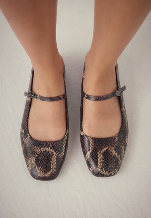 Piedi che indossano ballerine Mary Jane marroni e beige con motivo a pelle di serpente, punta quadrata e cinturini alla caviglia, in piedi su una superficie chiara.