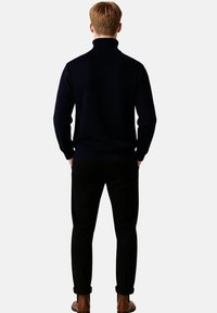 Námořnický rolák vyrobený z měkké tkaniny, přiléhavý střih, žebrované manžety, nošený s černými kalhotami, ukazující jednoduchou a čistou siluetu.