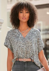 Blusa trasparente con motivo floreale in blu tenue, bianco e grigio, abbinata a una cintura intrecciata rosa. Maniche corte e scollatura a V.