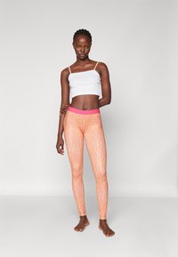 Rosa og oransje mønstrede leggings med en glatt overflate, høy midje, og en hvit cropped topp. Modellen står barfot og avslappet.