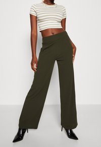 Olivgrüne, weit geschnittene Hose mit glatter Textur und hoher Taille, kombiniert mit einem gestreiften, cropped Oberteil und schwarzen spitzen Absätzen.