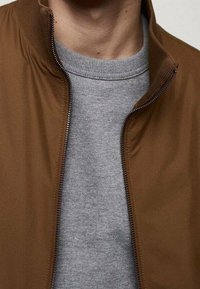 Veste marron zippée portée sur un pull gris à col rond. La veste présente un col montant, un design élégant et une texture lisse. Accent principal sur le détail de la fermeture éclair.