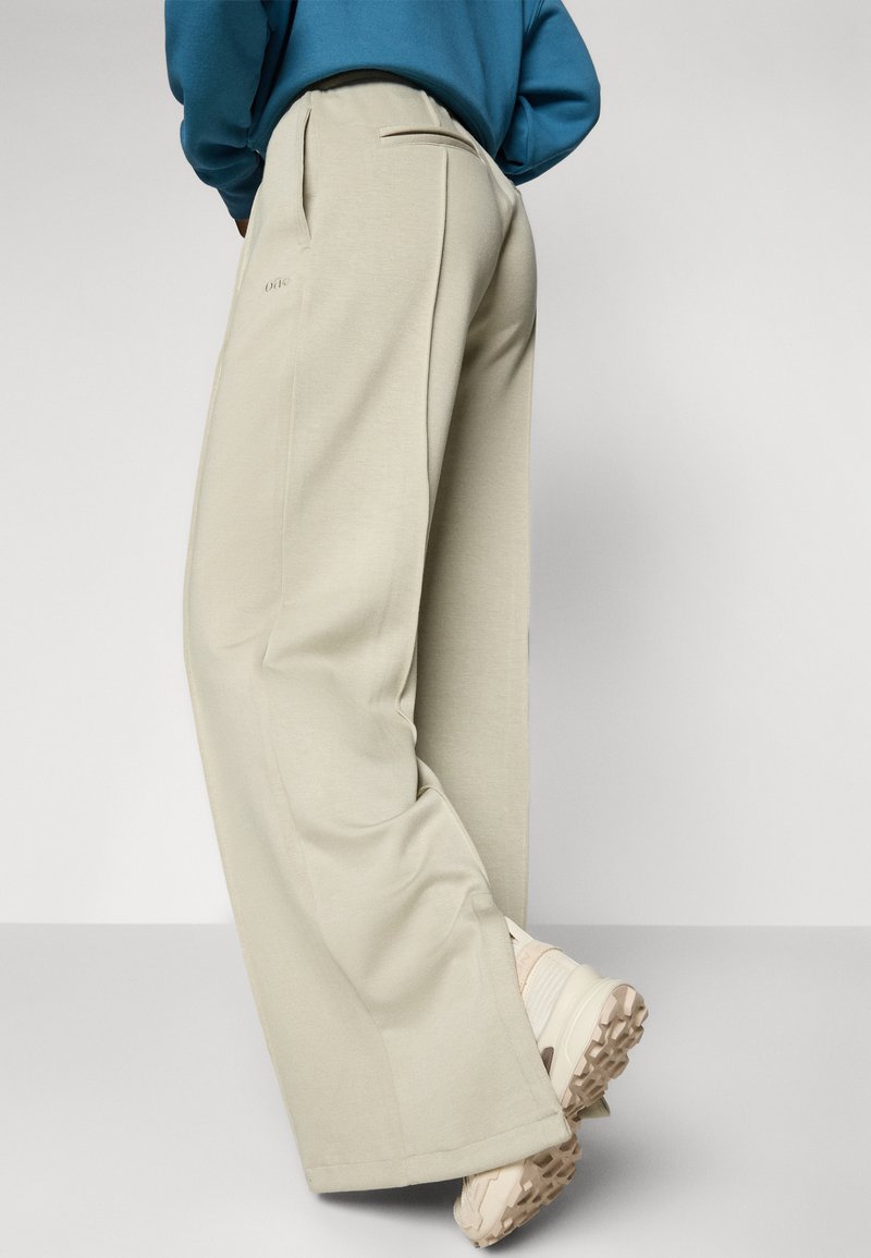 Pantaloni a gamba larga beige chiaro con una texture liscia e tasche laterali, abbinati a scarpe da ginnastica di colore chiaro e a una maglietta blu.