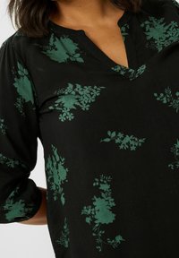 Sort bluse med grønne blomsterprint, med V-halsudskæring og 3/4 ærmer med lette manchetter. Blødt stof med en glat tekstur.