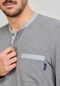 Hombre con barba corta que lleva una camisa henley gris con bolsillo en el pecho y tapeta de botones, ajustando un botón cerca del cuello.