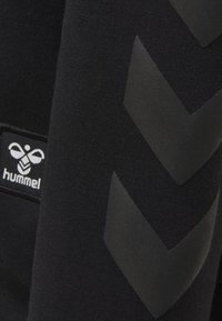 Svart sweatshirt med strukturerat material, med stora svarta chevronmönster och en vit logotagg märkt "hummel" på sidan.