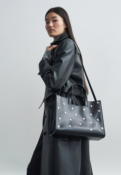 Zwarte leren totebag met zilveren studded details, twee handvatten aan de bovenkant en een afneembare schouderriem. Gladde textuur en gestructureerd ontwerp.
