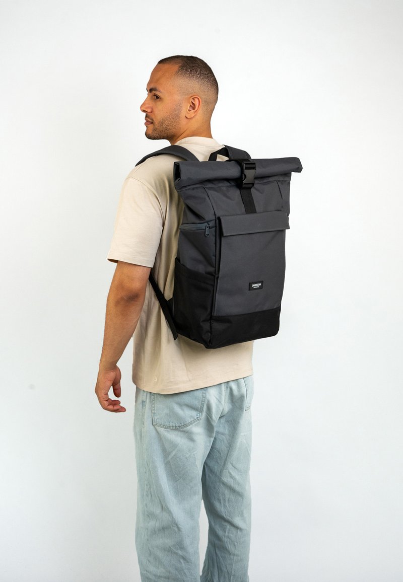 Black roll-top backpack with a front zip pocket, adjustable straps, and a textured fabric. Features a logo patch at the lower front.