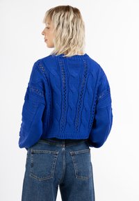 Pulover tricotat albastru, cu modele de cablu și cusături deschise. Design scurt, cu tiv striat, purtat cu blugi denim cu talie înaltă.