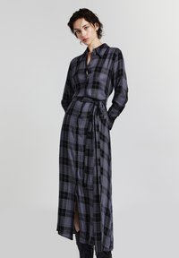 Donna che indossa un lungo vestito a quadri nero e grigio, con bottoni e un laccio in vita, in piedi con le mani dietro la schiena su uno sfondo semplice.