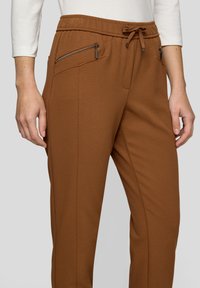 Braune Jogginghosen aus strukturiertem Stoff, mit einem Kordelzugbund und zwei Reißverschlusstaschen vorne. Eng an den Knöcheln zulaufend.