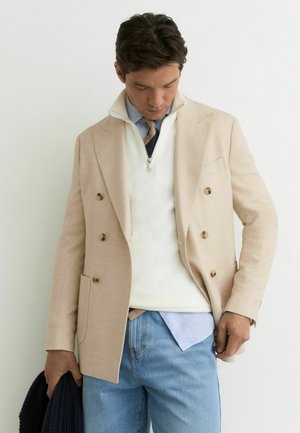 Mężczyzna w beżowej marynarce dwurzędowej, białym swetrze na suwak do połowy, niebieskiej koszuli, krawacie, jasnoniebieskich dżinsach, trzyma ciemny sweter, patrzy w dół.