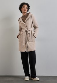 Manteau enveloppant en mélange de laine beige avec une capuche, une ceinture à nouer, deux poches avant et une fermeture à boutons. Porté sur un pull marron et un pantalon noir.