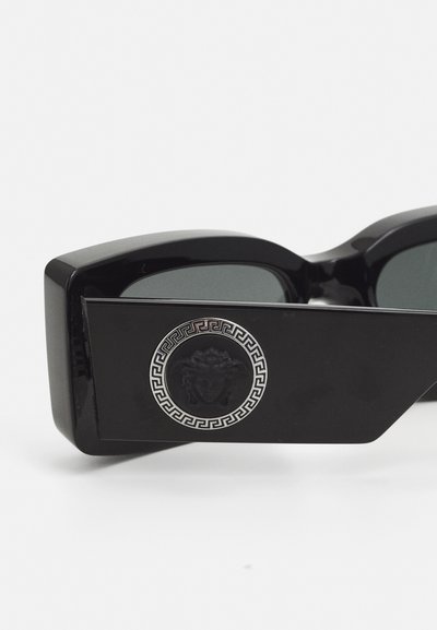Lunettes de soleil noires au design géométrique, ornées d'un emblème en métal représentant une Méduse et d'un motif de clé grecque sur le côté. Texture lisse.