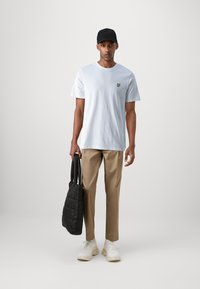 T-shirt di cotone azzurro chiaro con un piccolo logo giallo, abbinato a pantaloni beige, sneakers bianche e una borsa tote nera trapuntata.
