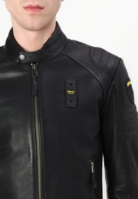 Giacca in pelle nera con colletto alto, chiusura con zip, spalline imbottite e una patch gialla con logo sul petto. Superficie a trama.