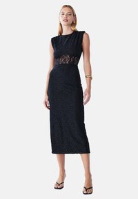 Robe en dentelle noire avec une silhouette ajustée, design sans manches, panneau transparent en dentelle à la taille et ourlet à hauteur des genoux. Accentuée par une quincaillerie minimale.