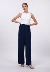 Bílý top bez rukávů v kombinaci s kalhotami s vysokým pasem ve navy barvě, které mají dva zlaté knoflíkové detaily a široké nohavice. Neutrální sandály.