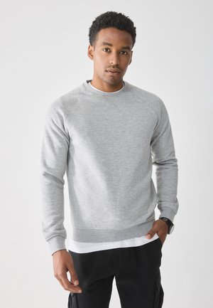 Man met kort, krullend haar, in een lichtgrijze sweatshirt, witte onderhemd, zwarte broek en een zwart horloge, staande tegen een witte achtergrond.