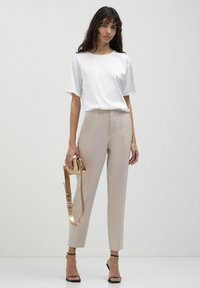 Top bianco a maniche corte con scollo rotondo, abbinato a pantaloni beige chiaro a vita alta. Il modello tiene una tracolla dorata. Sandali neri con tacco.