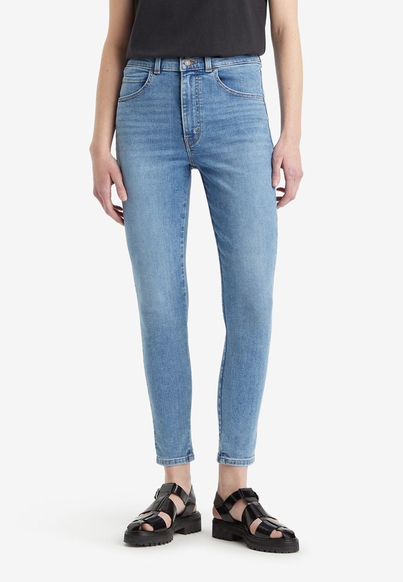 Personne portant un jean skinny bleu clair, un top noir sans manches et des sandales noires épaisses à bout ouvert, debout sur un fond blanc.