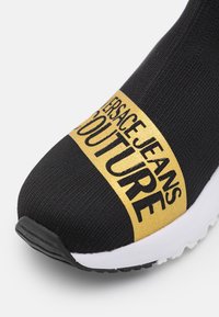 Svart stickad sneaker med ett guldlogo-band som säger "VERSACE JEANS COUTURE" och en vit gummisula. Strukturerad yta och elegant form.