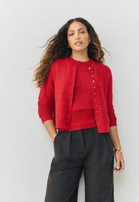 Red cardigan with heart-shaped buttons, round neckline, and long sleeves, paired with a matching red top and dark, loose-fit trousers.
