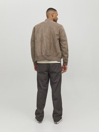 Giacca bomber in suede color taupe con polsini e orlo a costine. Abbinata a pantaloni grigi larghi e sneaker nere, vista da dietro.