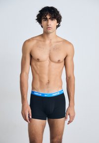 Boxer de Nike negros con una cinturilla elástica azul que muestra el nombre de la marca. El modelo tiene el pecho desnudo y el cabello corto y oscuro.