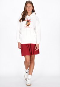 Fehér kapucnis pulóver ünnepi mackós grafikával és piros díszítésekkel, piros redőzött szoknyával és fehér tornacipővel párosítva.