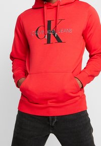 Punainen huppari pehmeää kangasta. Siinä on etutasku ja musta-hopea painettu "CALVIN KLEIN JEANS" -logo. Ilmataskut hihansuissa.
