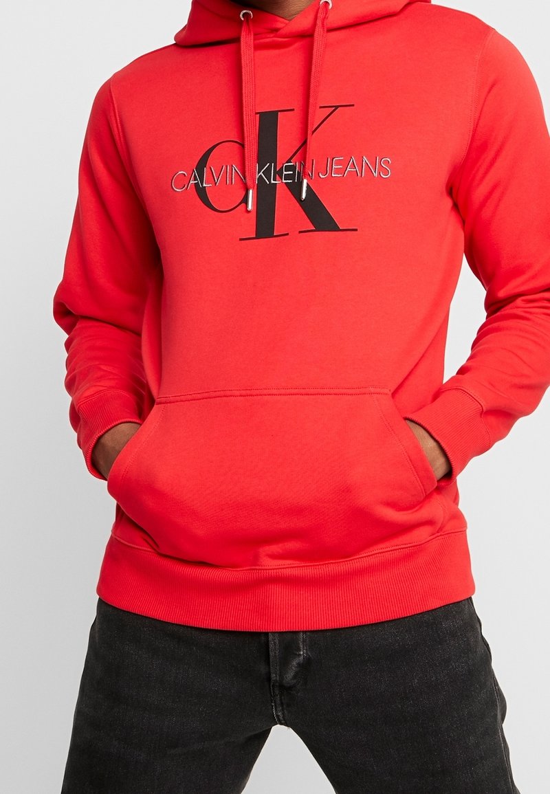 Punainen huppari pehmeää kangasta. Siinä on etutasku ja musta-hopea painettu "CALVIN KLEIN JEANS" -logo. Ilmataskut hihansuissa.