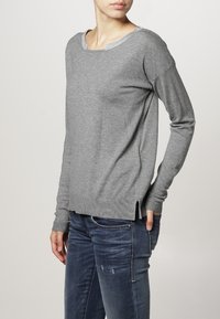 Grauer Langarmpullover mit rundem Ausschnitt und silbernem Besatz, mit seitlichen Schlitzen und schmalen Bündchen, kombiniert mit blauen Jeans.