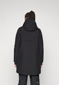 Osoba s pletenom kosom koja nosi crni zimski kaput s kapuljačom okrenuta je leđima, nasuprot neutralnoj sivoj pozadini. Odjeća izgleda funkcionalno i toplo.