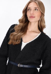 Zwarte, fuzzy cardigan met een diepe V-hals, gedragen over een witte top, getailleerd met een marineblauwe leren riem met een zilveren gesp.