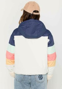 Farverig cropped jakke med en marineblå hætte, hvid krop og vandrette striber i mint, pink og fersken. Struktureret stof og tætsiddende pasform.