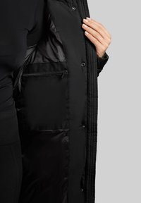 Juoda izoliuota striukė su siuvinėta išorine dalimi, turinti lygią, blizgią vidinę pamušalą ir užtraukiamą kišenę viduje.