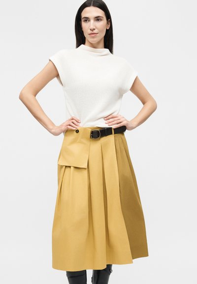 Γυναίκα με μακριά σκούρα μαλλιά που φοράει λευκό πλεκτό τοπ με κοντά μανίκια και πιετέ φούστα μουσταρδί χρώματος με μαύρη ζώνη και τσαντάκι.