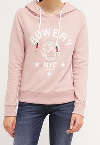 Person som bär en rosa hoodie med "BOWERY NYC" och en tiger-grafik, tillsammans med blå jeans mot en enfärgad bakgrund.