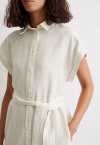 Abito camicia in lino crema con maniche corte, apertura frontale con bottoni e cintura legata in vita. Texture liscia con un design casual.