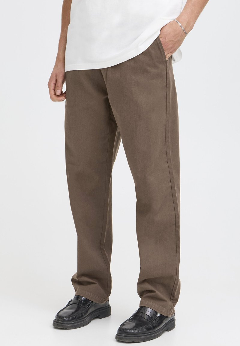 Pantaloni di cotone marrone con taglio dritto, tasche laterali visibili e trama sottile. Abbinati a calzature nere con finitura liscia.