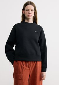 Νεαρή γυναίκα με μακριά καστανά μαλλιά, φοράει μαύρο φούτερ με λογότυπο και καφέ cargo παντελόνι, σε απλό λευκό φόντο.