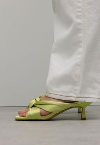 Groene satijnen sandalen met hak en een geknoopt ontwerp. Heeft een lage stiletto-hak en een open teen. Gepaard met witte, iets uitlopende broeken.