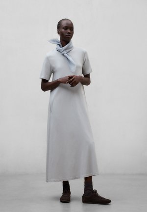 Kobieta w jasnoszarej, krótkorękawowej sukience maxi, z jasnoniebieskim szalikiem na szyi, ciemnych skarpetkach i brązowych butach, stojąca na tle jednolitej ściany.