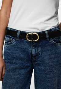 Melns ādas josta ar zelta ovālu sprādzi, nēsāta pār zilām džinsa biksēm, ar izbalējušu tekstūru un klasisku kabatas dizainu.